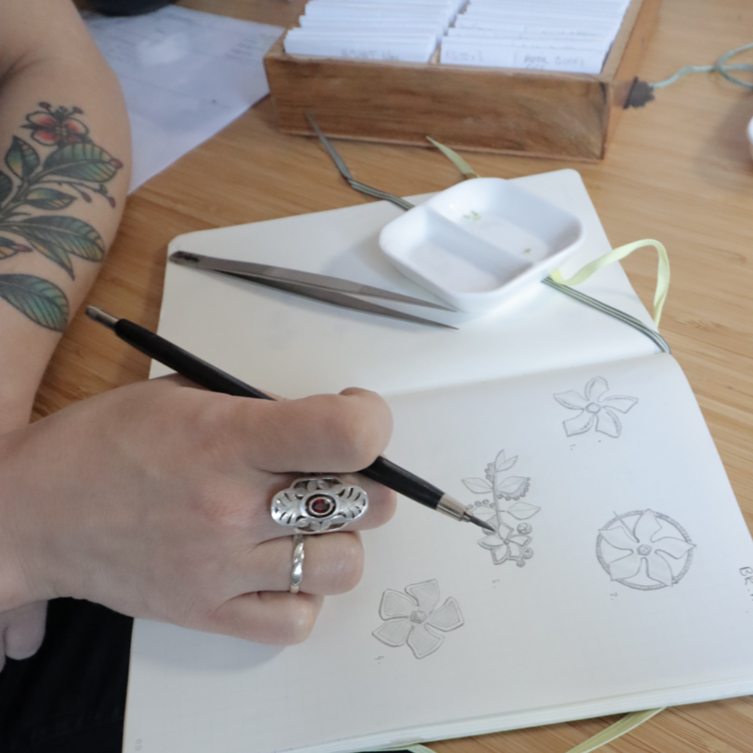 A picture of a hand drawing floral motives for jewellery and wearing two silver rings, one with a red garnet gemstond. Casez Jewellery.