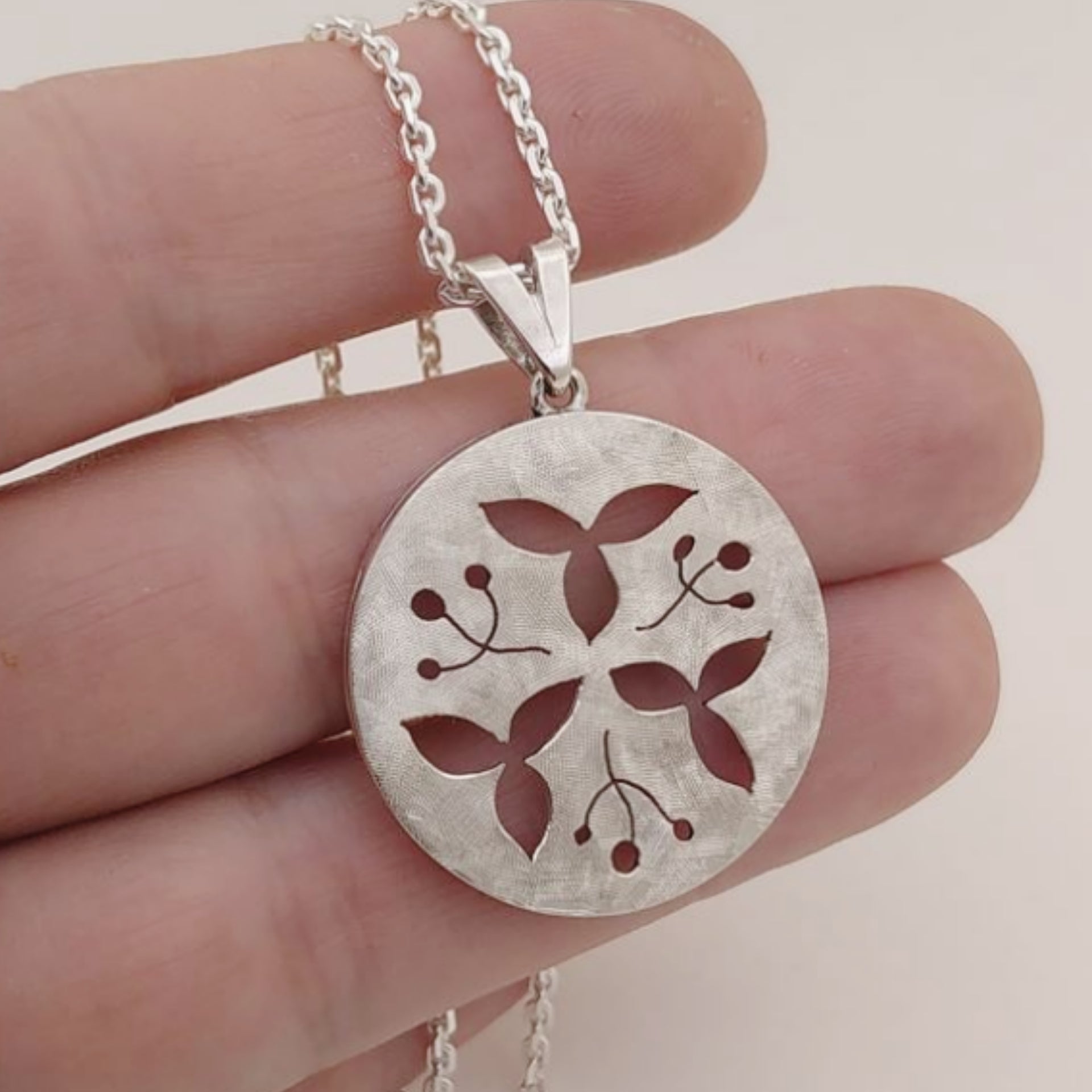 A round silver pendant in which floral and leave patterns are cut. Made by hand by Casez Jewellery.