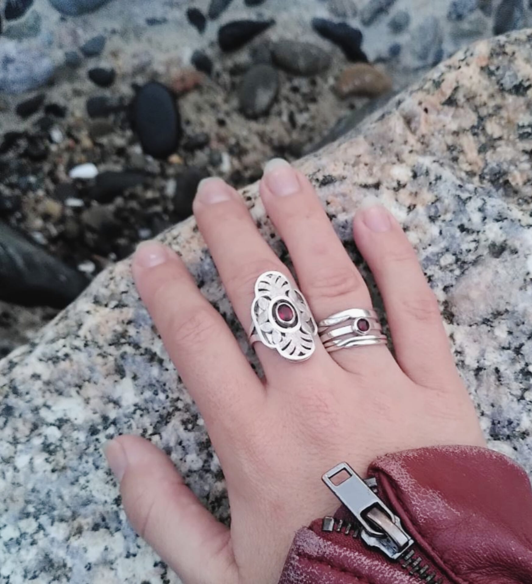 A hand ouside on a rock, wearing four silver rings. Two of these rings display red garnet gemstones. Made by hand by Casez Jewellery.