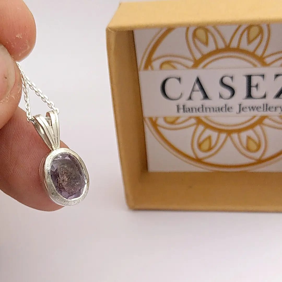 A silver pendant displaying an amethyst in oval cut. A cat claw and cat hairs are hidden below the stone. Made by hand by Casez Jewellery.
