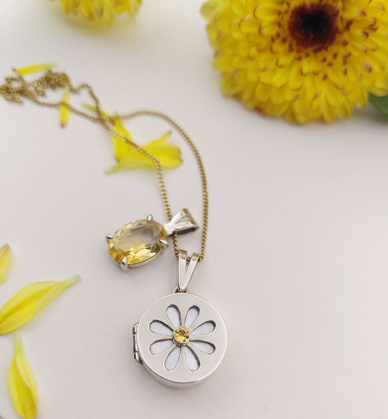 A small locket pendant displaying a daisy flower pattern on top, with a citrin and mother of pearl. Is attached on a gold chain together with another pendant, a simple citrin in oval cut. Made by hand by Casez Jewellery. 