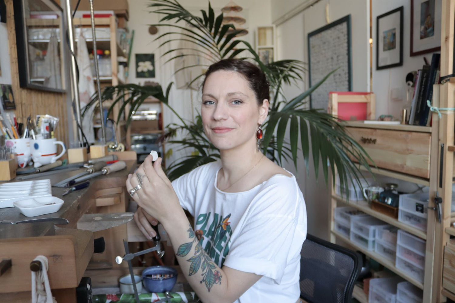 A portrait of Marion Chartier, from Casez Jewellery, in her workshop. She holds a piece of silver in her hand and wears a greenpeace tshirt and a pair of earrings with large baltic amber cabochons. 